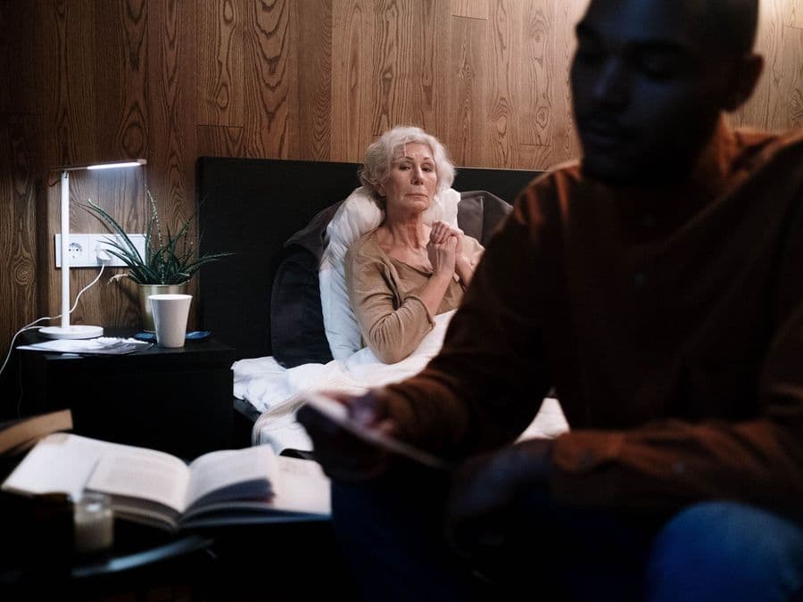 A care worker sitting beside an older woman in a softly lit room late in the evening.