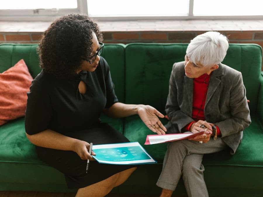 A care worker and an older woman sitting together on a sofa, looking at something and talking.