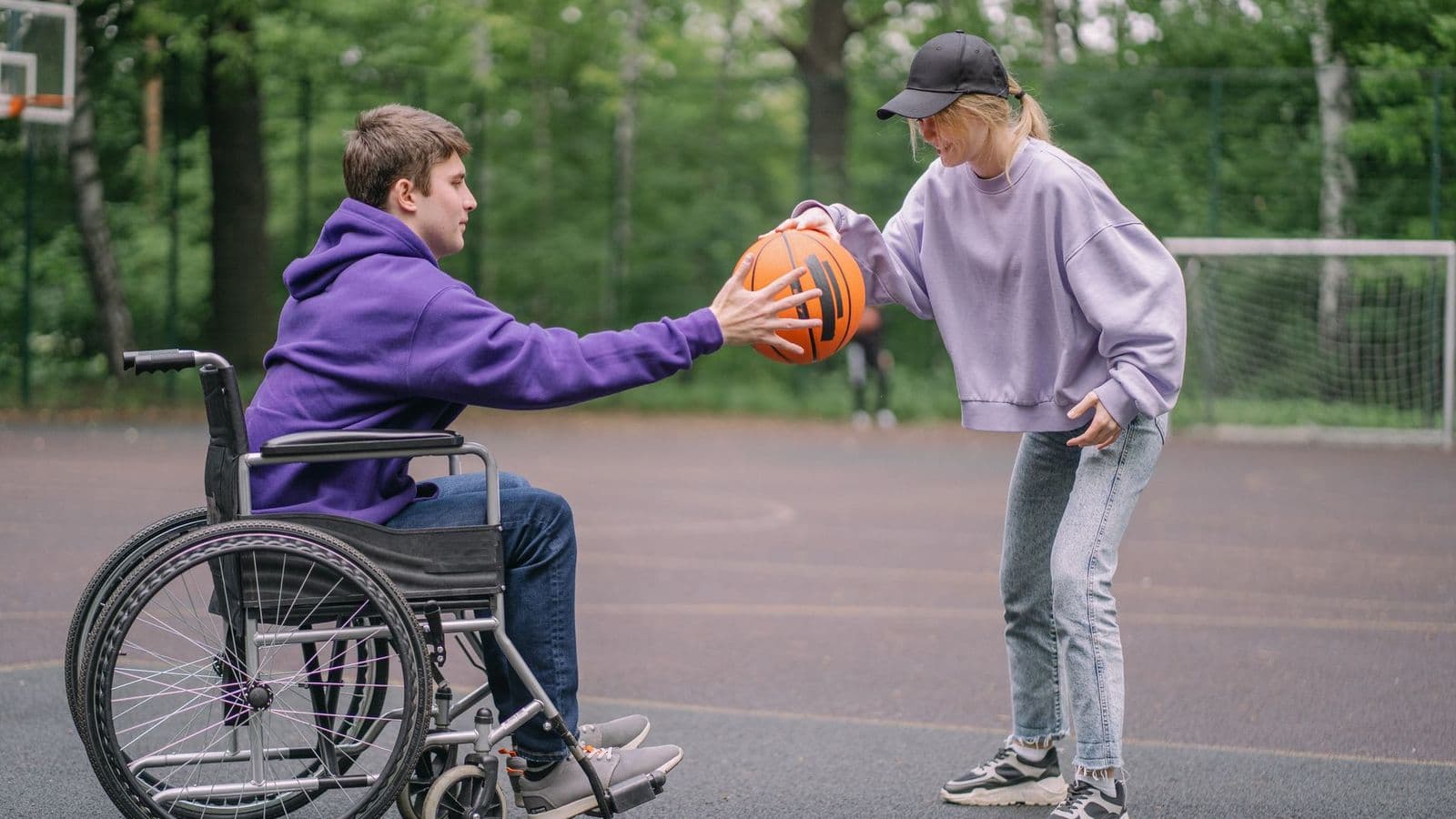 A young man using a wheelchair playing basketball outdoors with a support worker passing him the ball.