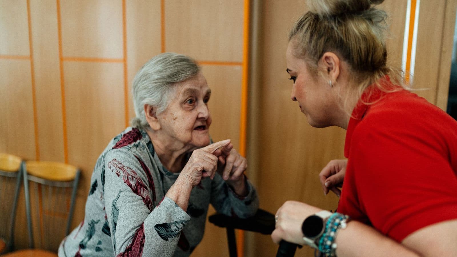 A care worker sitting close to an older woman in her own living room, helping her with a hand exercise.