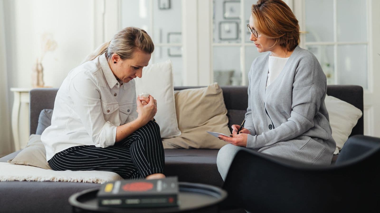 A counsellor and a client sitting together on a sofa, talking while the counsellor takes notes.