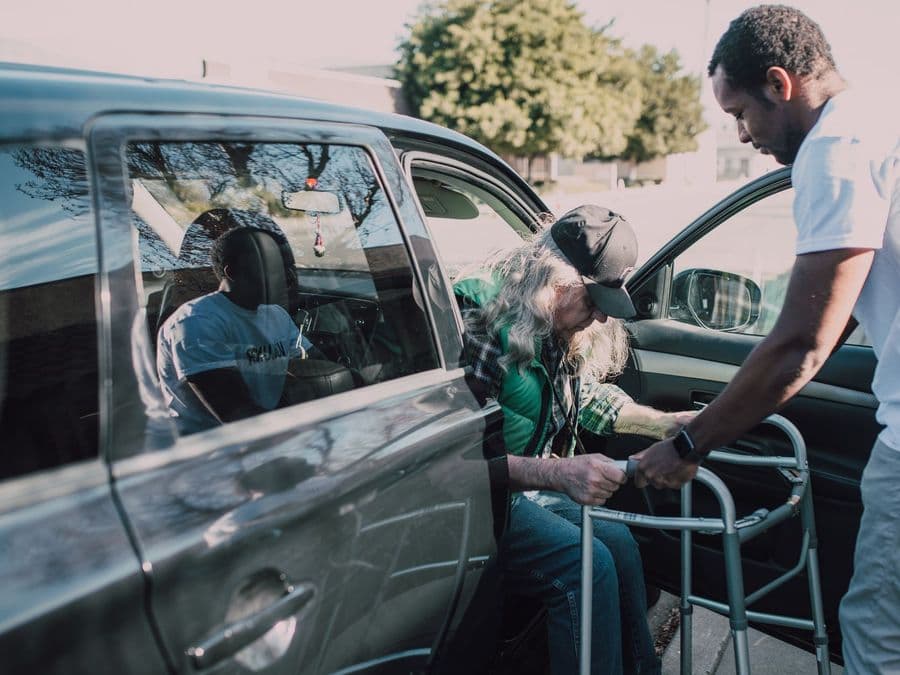 A care worker helping an older person with a walking frame get into a car.